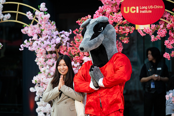 Woman posing with a costumed anteater character in red attire, hands together in greeting, in front of pink blossoms and a “UCI Long US-China Institute” sign.